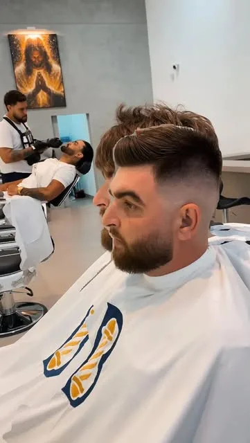 Cliente com degradê alto e barba cheia, de capa branca, sentado no salão