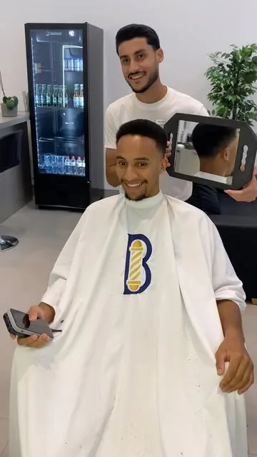 Cliente sorrindo enquanto confere o corte num espelho de mão, com o barbeiro atrás